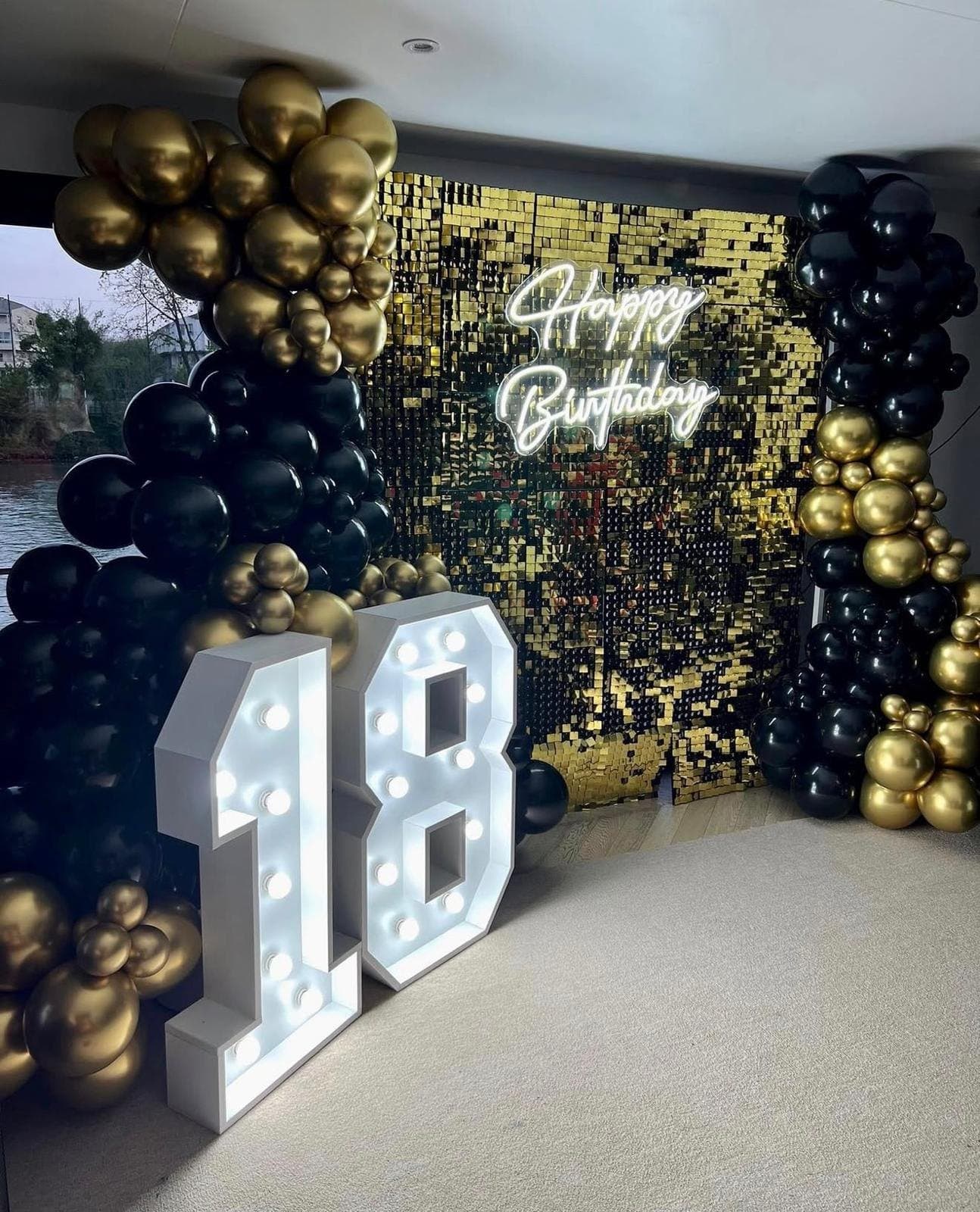 Chiffres lumineux géants 18 devant un fond à paillettes dorées et des ballons noirs et or